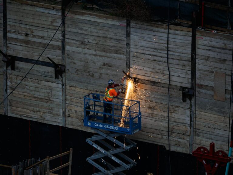 A construction worker welds on a raised platform, creating sparks on an industrial site.
